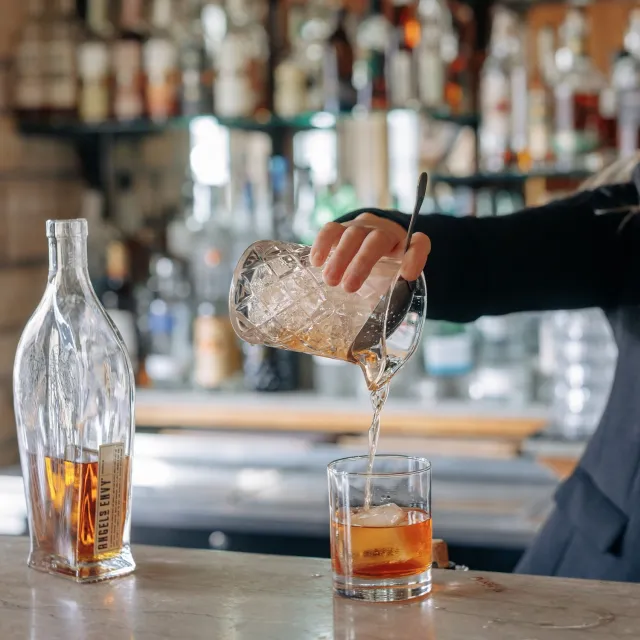 A bartender pours whiskey over ice into a glass, with a decorative bottle in the foreground and a blurred bar shelf in the background.