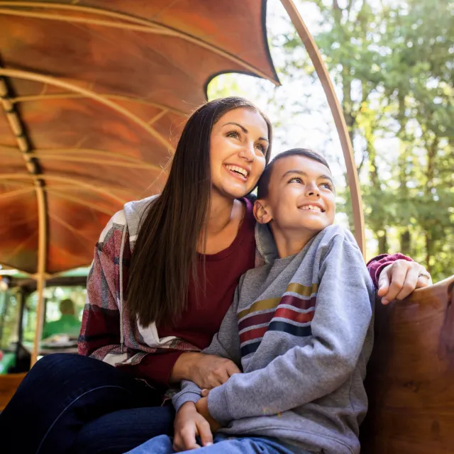 A woman and a child sit closely together on a wooden bench illuminated by soft sunlight, surrounded by lush greenery.