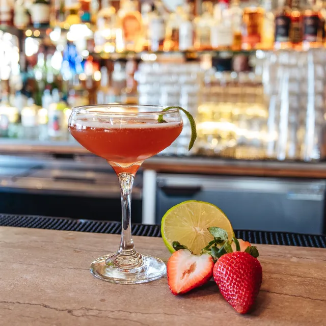A vibrant cocktail with a lime twist sits on a bar, alongside fresh strawberries and lime slices, against a blurred backdrop of bottles.