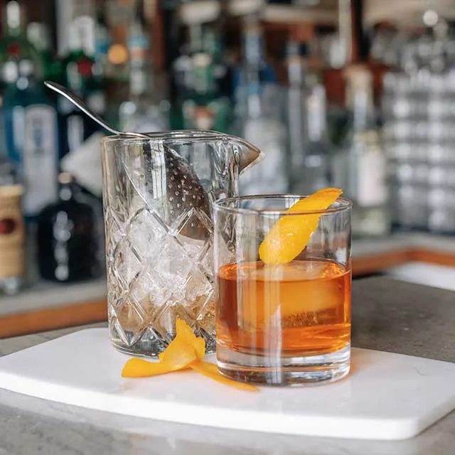 A polished glass with a cocktail and orange twist sits beside a mixing glass filled with ice, against a backdrop of liquor bottles.