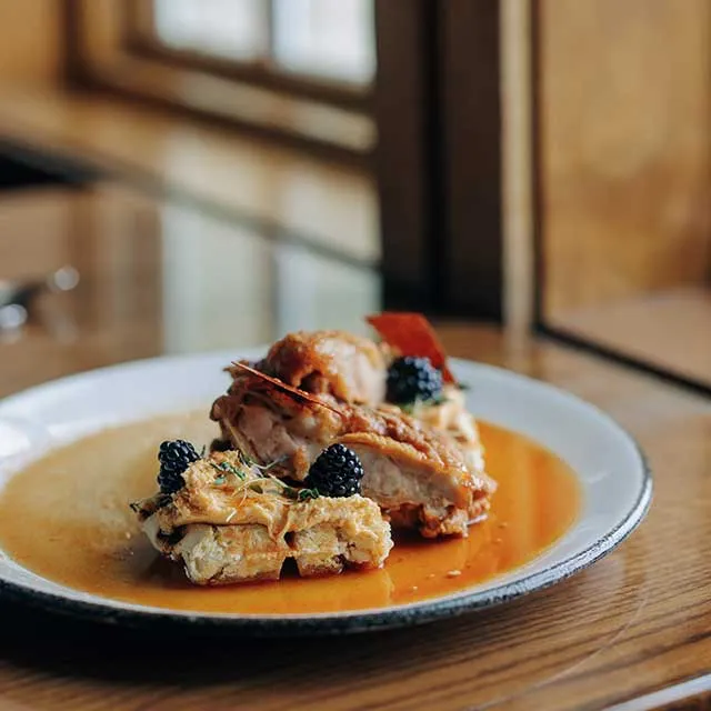 A plated dish featuring succulent chicken atop a crispy waffle, garnished with blackberries and a rich sauce.