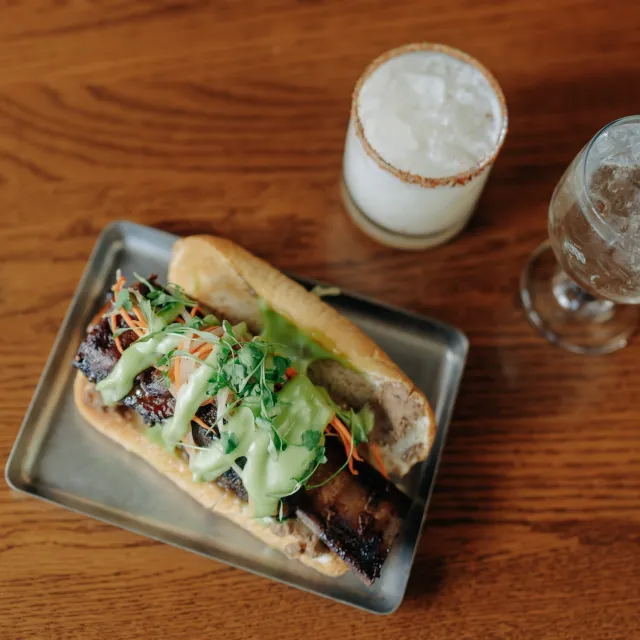 A grilled meat sandwich topped with green sauce, cilantro, and veggies, served on a metal tray with two drinks in the background.