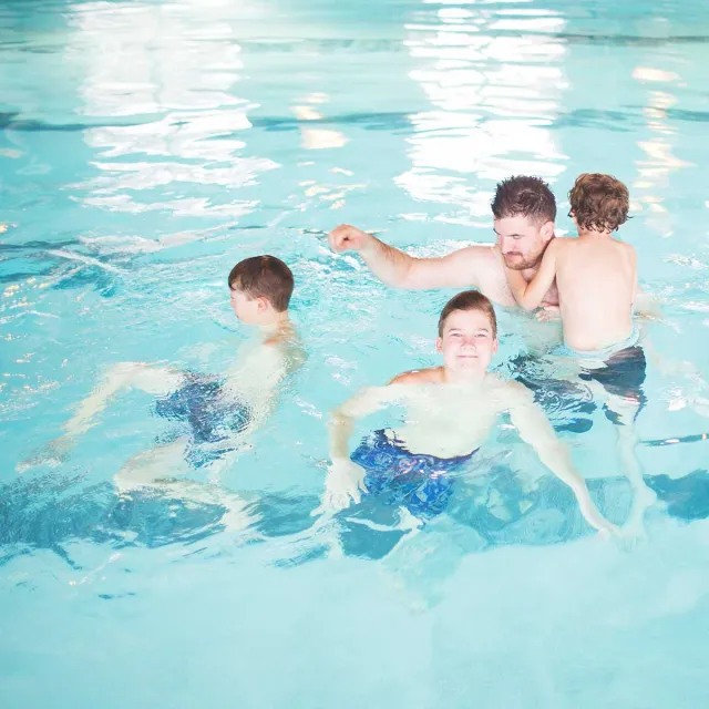 A group of children play and swim in a bright, clear indoor pool, enjoying splashes and laughter together.