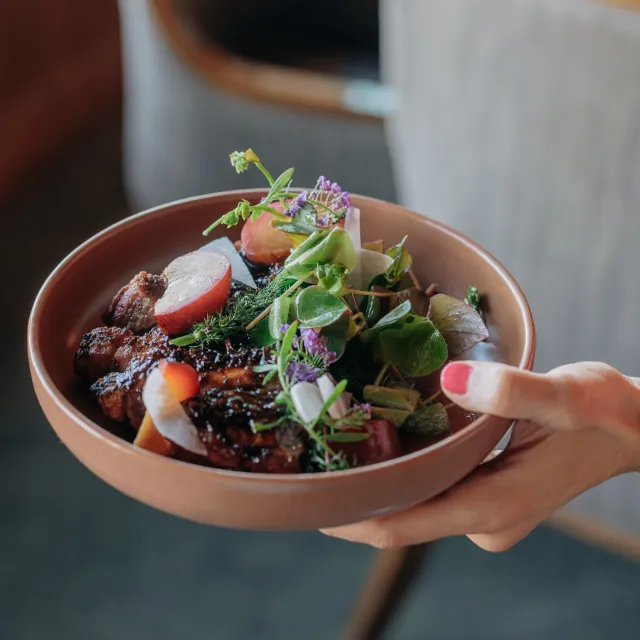 A hand holds a brown bowl filled with grilled meat, colorful vegetables, and fresh herbs, presented elegantly.