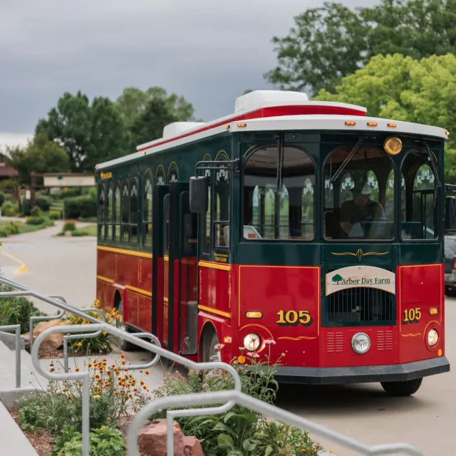 A colorful red and green trolley labeled "Arbor Day Farm" drives along a landscaped path with flowers and greenery.
