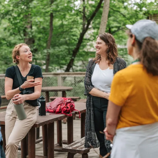 A small group of people engage in conversation outdoors, surrounded by trees, with a picnic table and backpacks nearby.