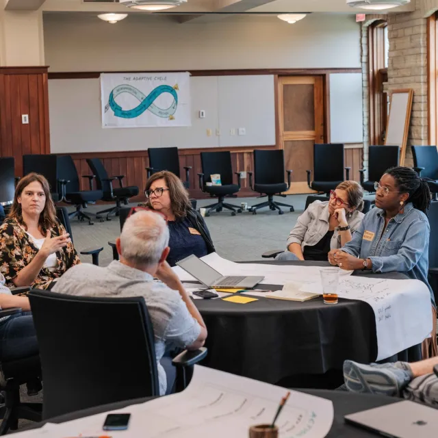 A group of individuals engaged in a discussion around a table, with presentation materials and a chart on the wall in a professional setting.