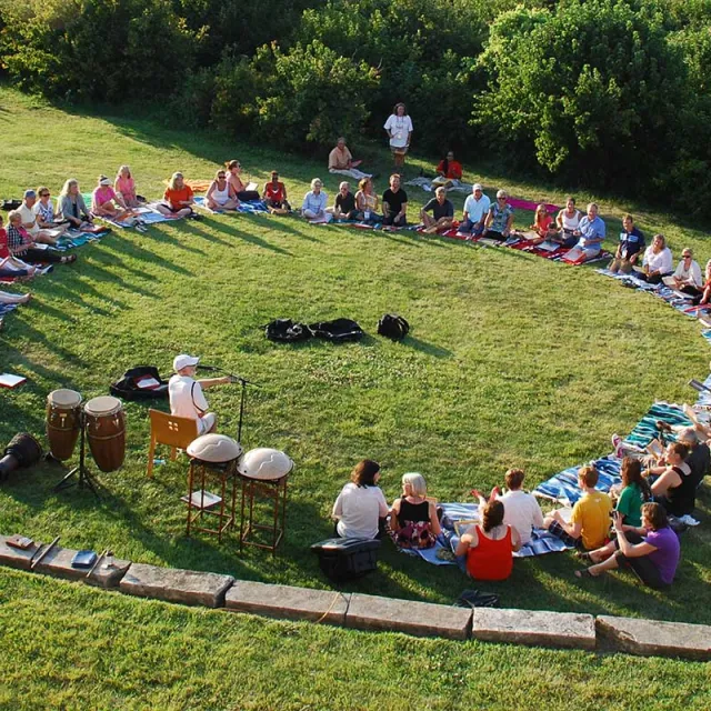 A group of seated people in a circle on the grass, engaged in a music or meditation session, with instruments nearby.