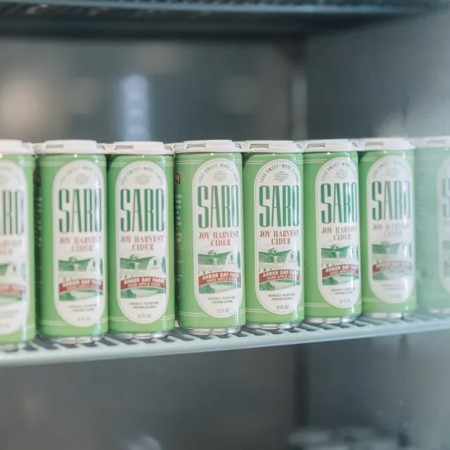 A refrigerator shelf filled with cans of Saro Joy Harvest Cider, featuring green packaging with bold text and scenic graphics.