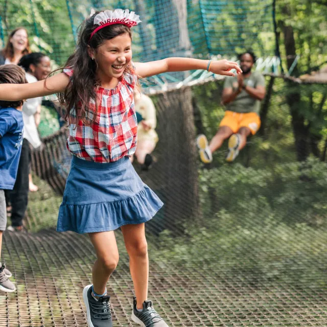 A child in a checked shirt and denim skirt balances on a rope bridge, enjoying a playful moment among friends in a vibrant outdoor setting.