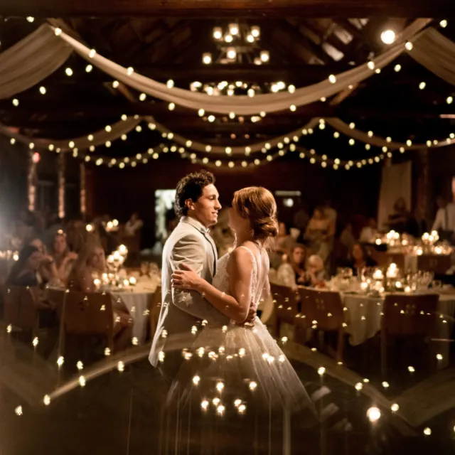 A couple dances romantically under soft, twinkling lights at a beautifully decorated wedding reception. Guests enjoy the enchanting atmosphere.