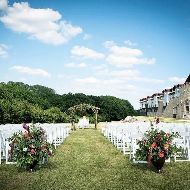 A picturesque outdoor wedding setup with white chairs, a floral arch, and lush greenery under a clear blue sky.