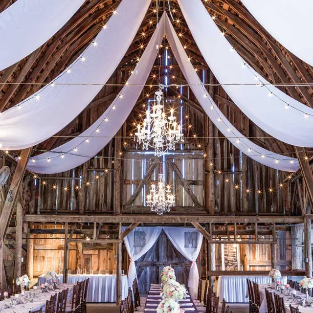Elegant barn interior decorated for a wedding, featuring draped fabric, sparkling chandeliers, and floral centerpieces along long tables.