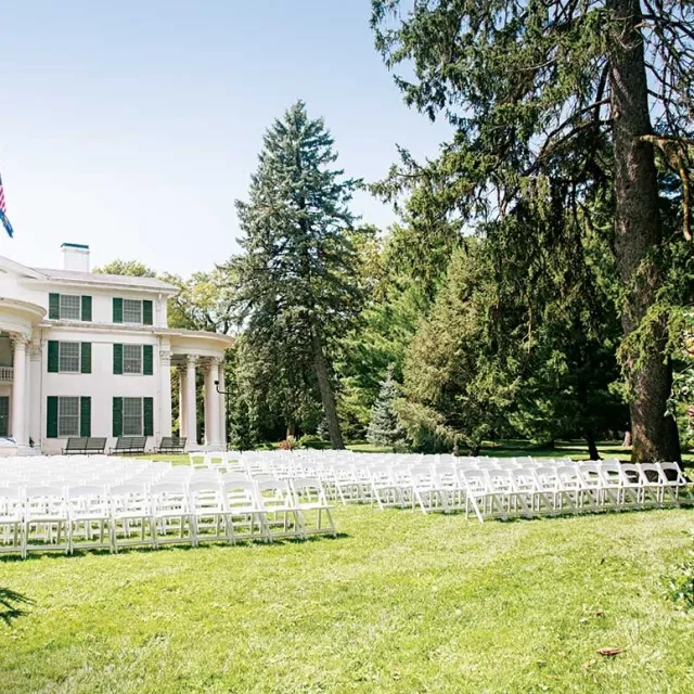A grand white mansion with green shutters in a scenic park, featuring rows of white chairs set up for an outdoor event.