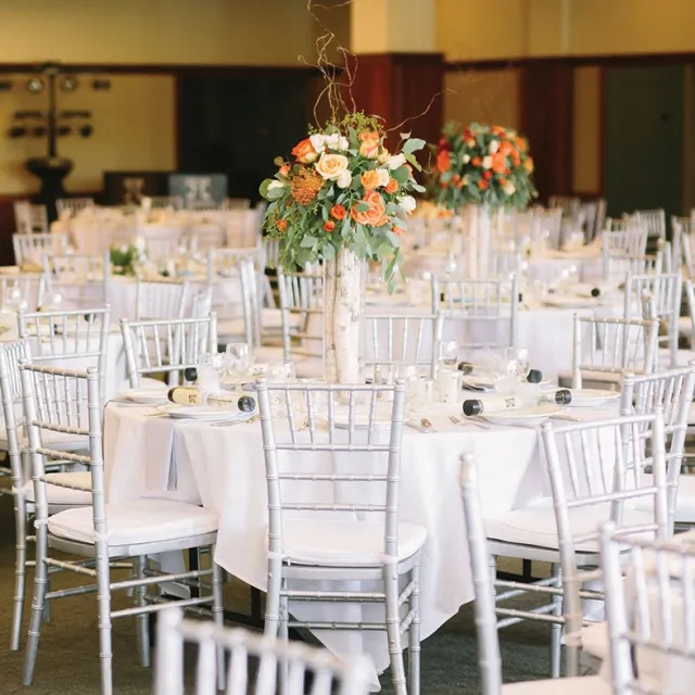 Elegant dining setup featuring silver chiavari chairs and tables adorned with floral centerpieces in warm tones, ready for an event.