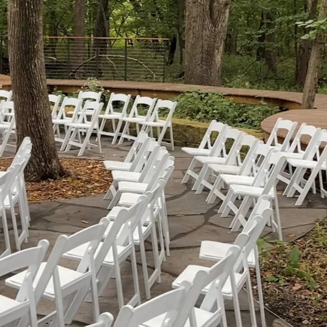 A scenic outdoor setting featuring rows of white folding chairs arranged in a circular stone area, surrounded by trees and greenery.
