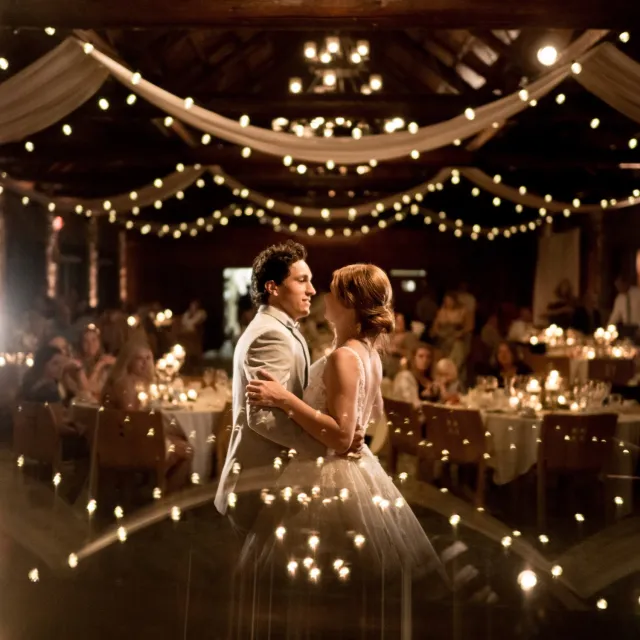 A couple dances romantically under soft, glowing lights at a beautifully decorated wedding reception, surrounded by seated guests.