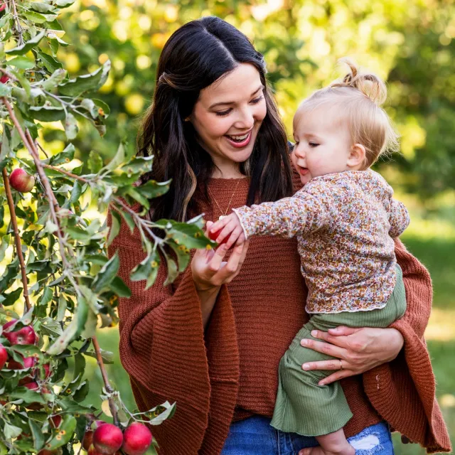 A woman holds a toddler in an apple orchard, both reaching for ripe red apples among green leaves.