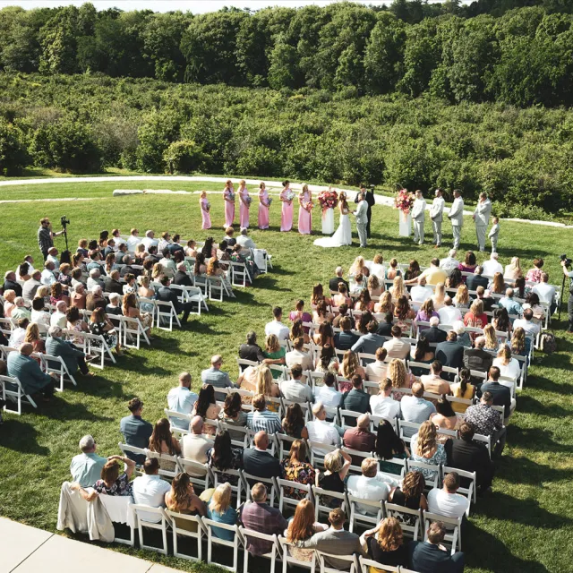 A large outdoor wedding ceremony with guests seated on white chairs, bridal party in pink dresses, and lush greenery in the background.