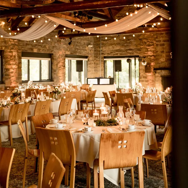 Elegant banquet hall with wooden tables set for dining, adorned with candles and greenery, under warm lights and draped fabric.