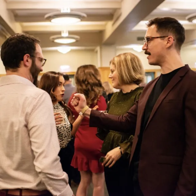 A group of people engaged in conversation at a lively event, with drinks in hand and festive attire in a warmly lit venue.