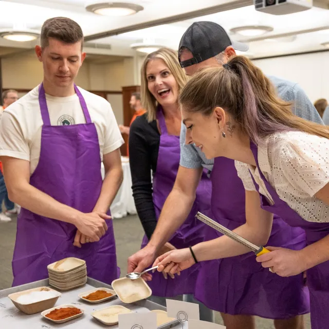 A group of people in purple aprons is gathered around a table measuring spices with various bowls laid out in front of them.