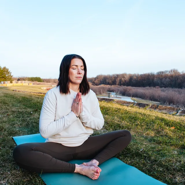 A person sits cross-legged on a yoga mat outdoors, with hands in a prayer position, surrounded by grass and trees.