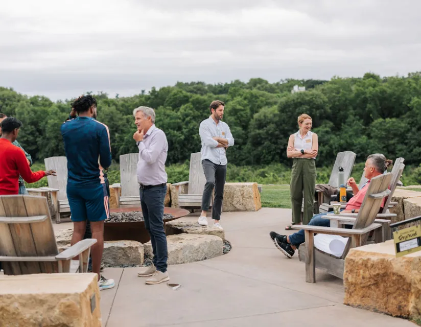 A group of six people engage in conversation around a fire pit, surrounded by greenery and wooden chairs on a cloudy day.