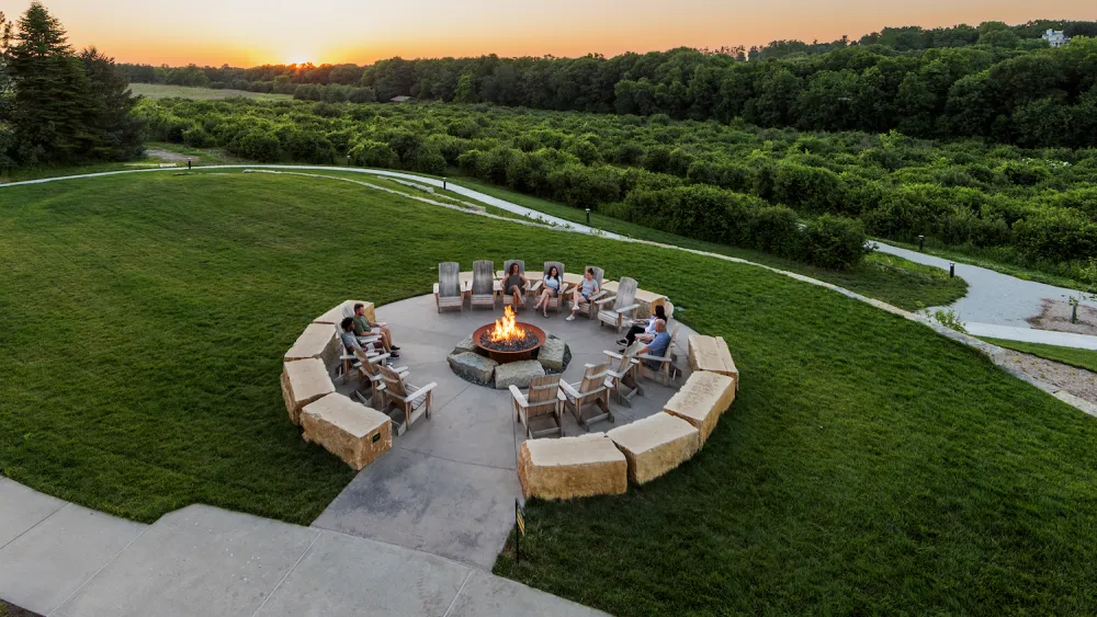 A group of people sit around a large circular fire pit on a grassy lawn with trees and a setting sun in the background.