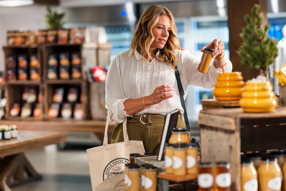 A woman inspects a jar of honey at a market stall filled with various jars and bottles of preserves and honey products.