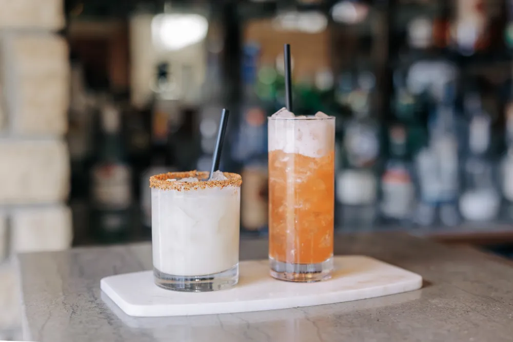 Two cocktails on a white serving board: a creamy, garnished drink and a vibrant orange beverage with ice, set on a marble table.