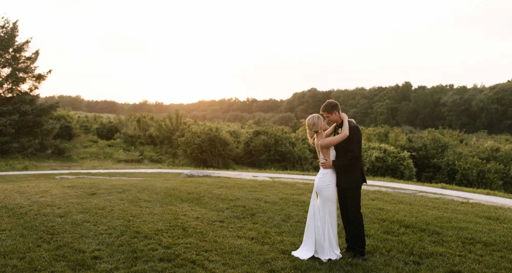 A couple embraces in a grassy field at sunset, surrounded by lush greenery and a soft, glowing sky.
