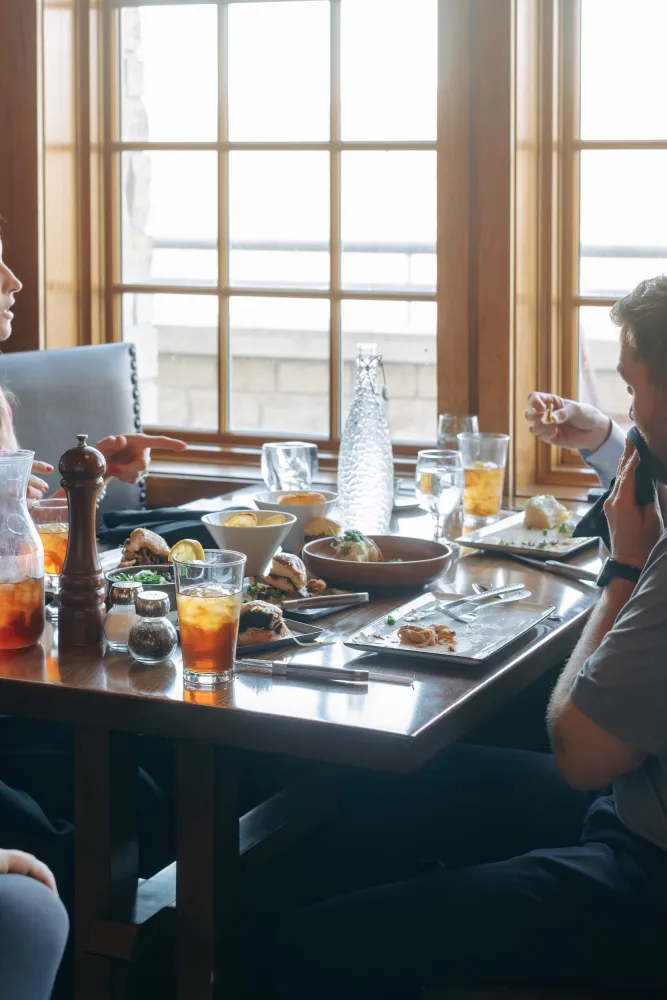 A wooden dining table filled with dishes and drinks, with friends engaged in conversation by a sunny window.