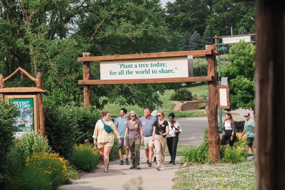 A diverse group of people walks along a path, passing under a wooden sign that reads, "Plant a tree today, for all the world to share."