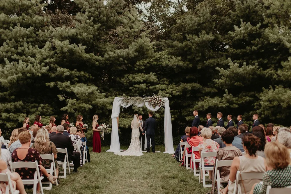 Outdoor wedding ceremony with guests seated on either side of an aisle and the couple stands under a floral archway surrounded by trees.