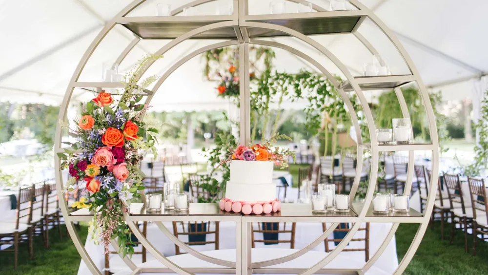 A beautifully decorated cake stand featuring a white wedding cake with floral accents, surrounded by vibrant flowers and candles under a tent.