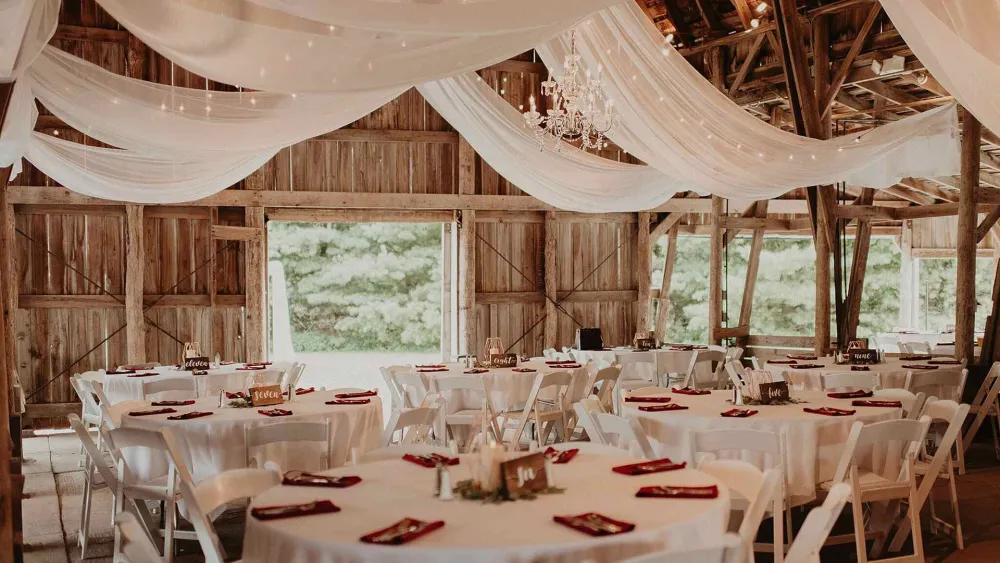 Elegant barn interior decorated for an event, featuring draped fabric, a chandelier, round tables with white linens, and red napkins.