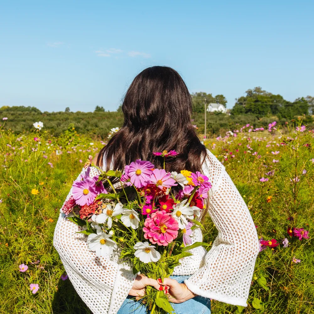 A woman in a white sweater and jeans holds vibrant pink, purple, and white flowers behind her back, standing in a colorful wildflower field under a clear blue sky.