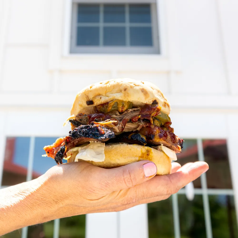 A hand holds a towering sandwich filled with smoked meat, pickles, and sauces, set against a light, modern building backdrop.