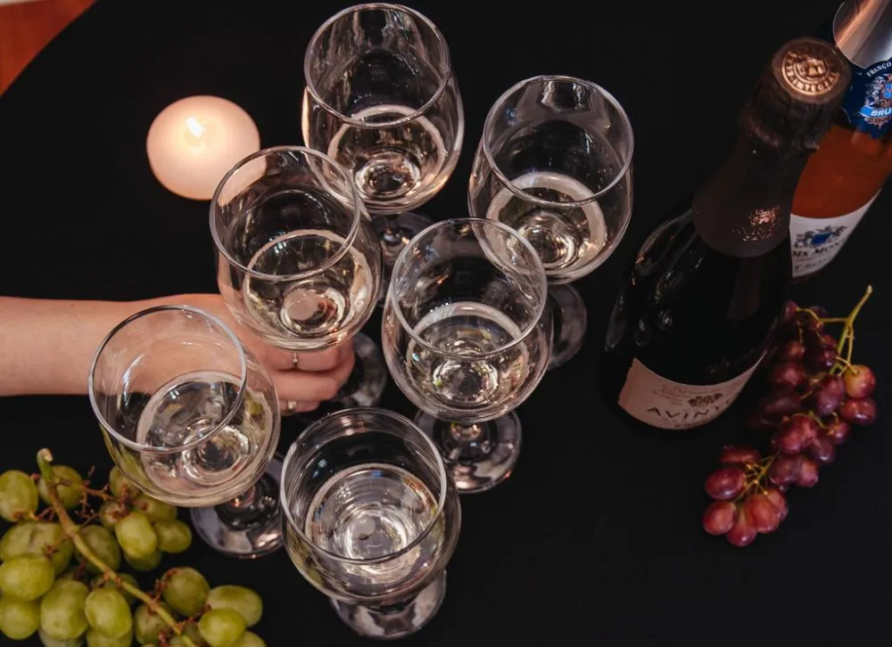 Six wine glasses filled with white wine, a bottle of sparkling wine, grapes, and a lit candle on a black tablecloth.