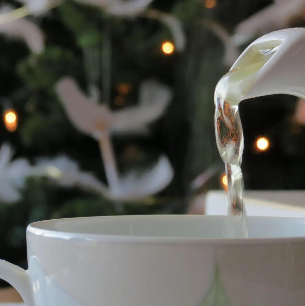 A teapot pours steaming tea into a white cup, set against a softly lit background of a decorated Christmas tree.