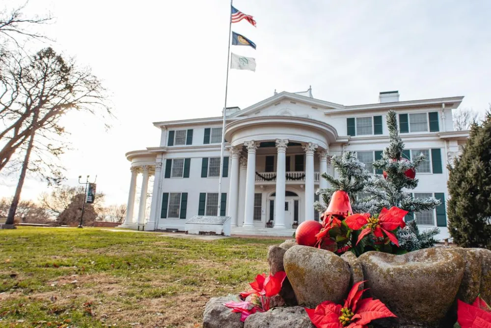 Historic white mansion with green shutters, flags flying, adorned with festive decorations and a small Christmas tree in the foreground.