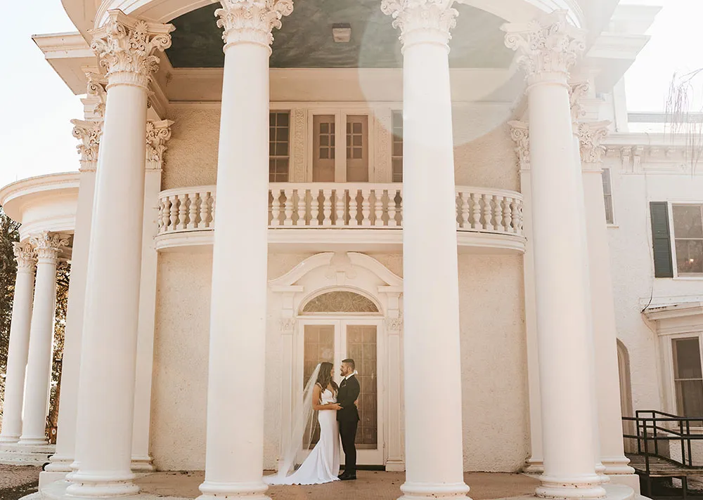 A bride and groom share a tender moment in front of an elegant, columned mansion on their wedding day.