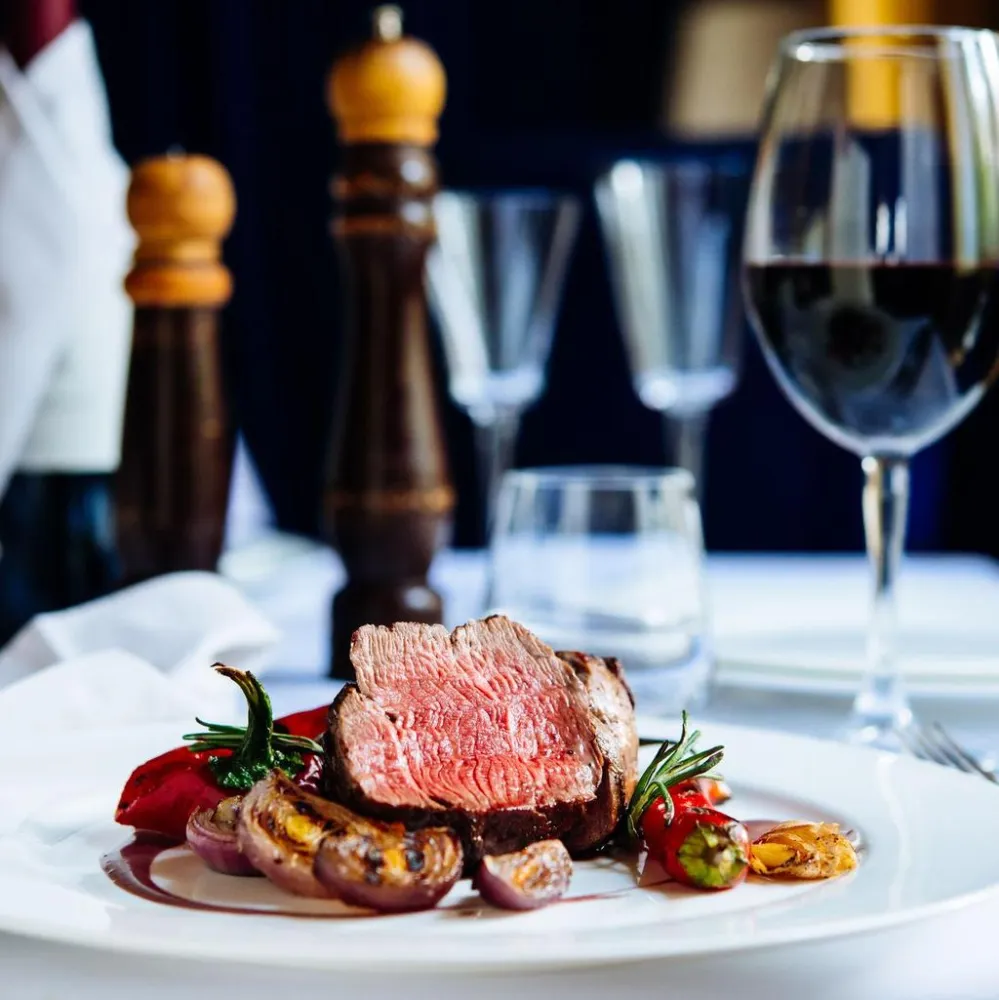 A gourmet dish of medium-rare steak garnished with grilled vegetables on a white plate. A glass of red wine and pepper mills create an elegant dining setting.