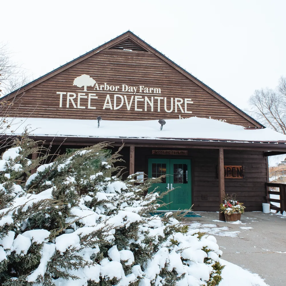 A wooden building with a sign reading "Arbor Day Farm Tree Adventure," surrounded by snow-covered bushes in winter. The doors are green and open.