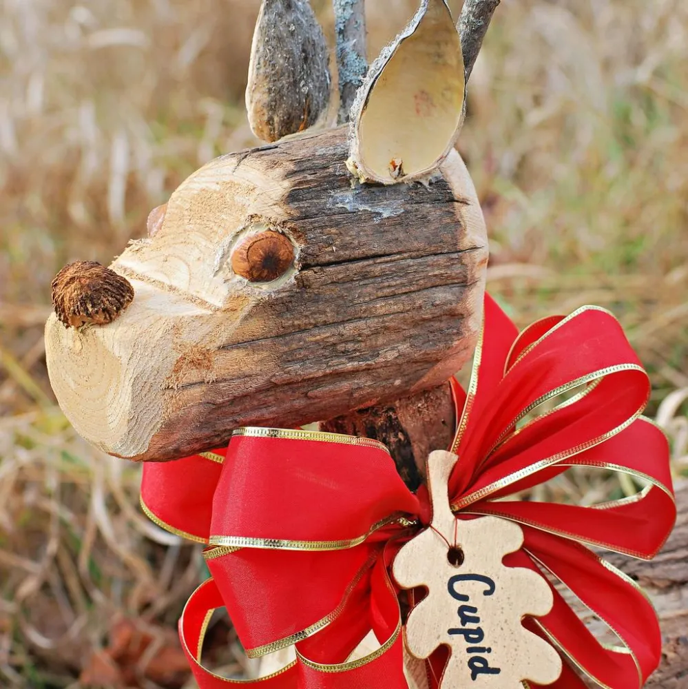 Close-up of a wooden reindeer figure adorned with a red bow and gold trim. A leaf-shaped tag labeled "Cupid" hangs from the bow.