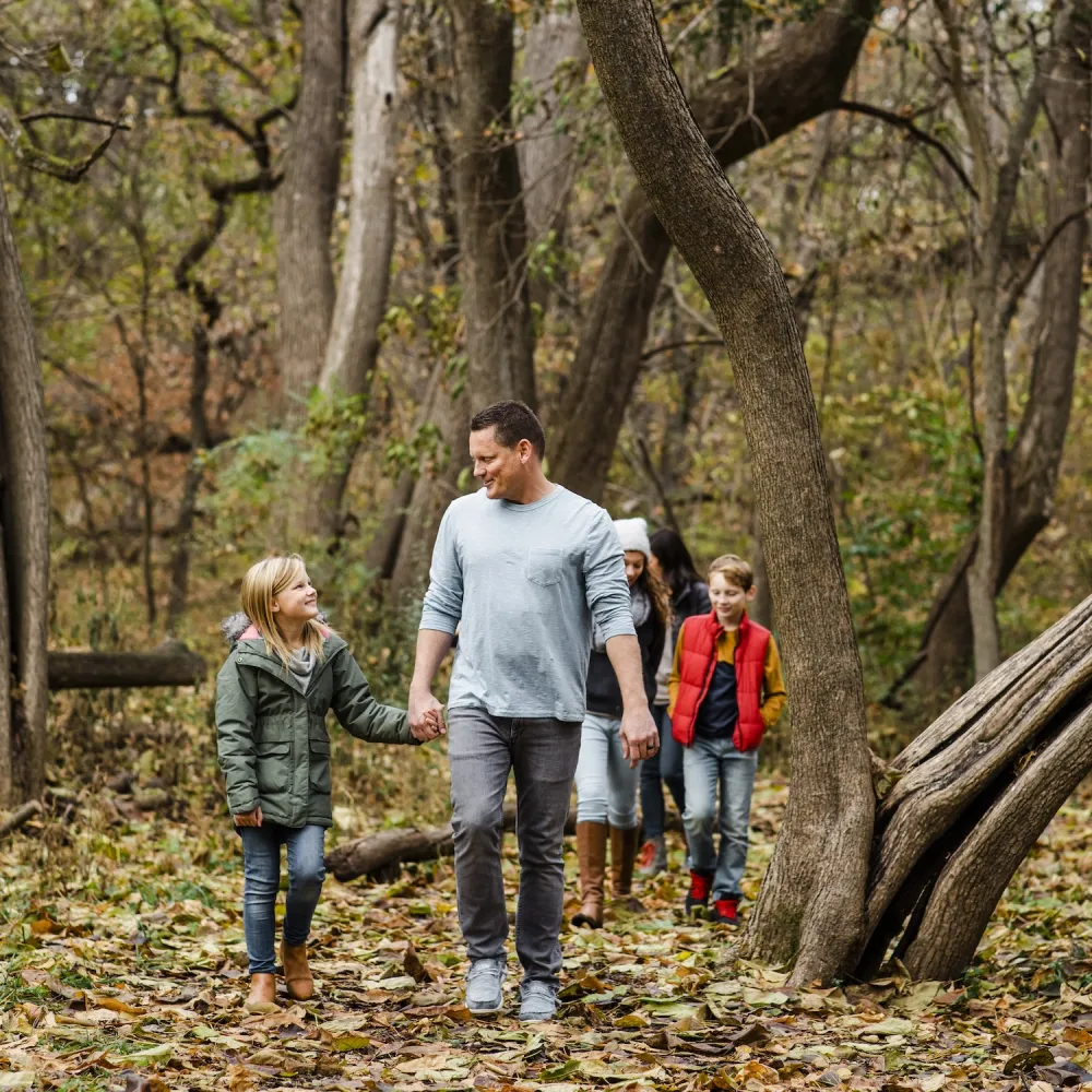 A man and four children stroll through a serene forest, surrounded by autumn foliage and tall trees.