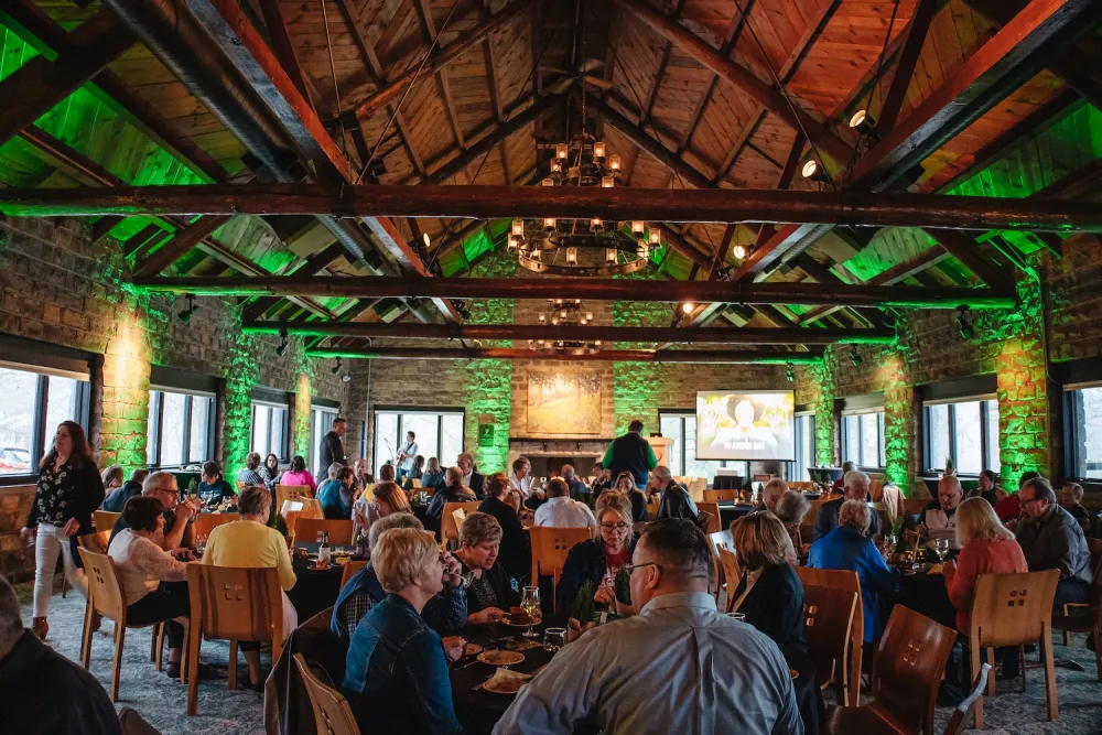 A lively restaurant interior with wooden beams, green lighting, and tables filled with guests enjoying food and drinks. A screen displays content.
