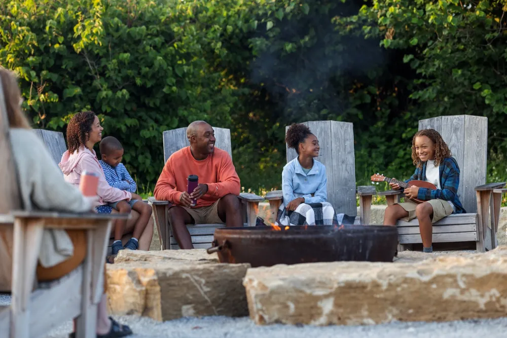 A group of people sit around a fire pit, enjoying a cozy outdoor gathering amidst lush greenery, with some playing music.