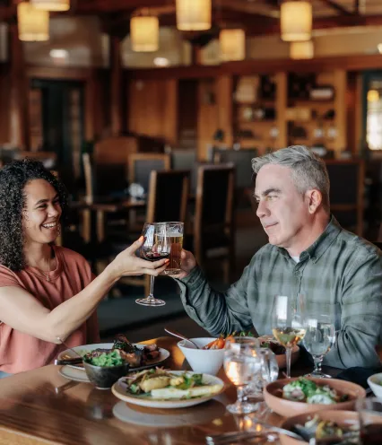 Two people toast with glasses in a cozy restaurant setting, surrounded by plates of food and soft lighting.